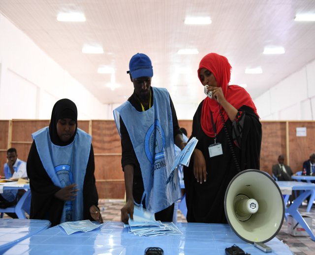 Eine Gruppe von Menschen versammelt sich um einen Tisch mit einem Megafon, Papieren und anderen Gegenständen, die scheinbar eine Wahllokal sind, mit anderen im Hintergrund unter beleuchteten Deckeninstallationen sitzend und stehend.
