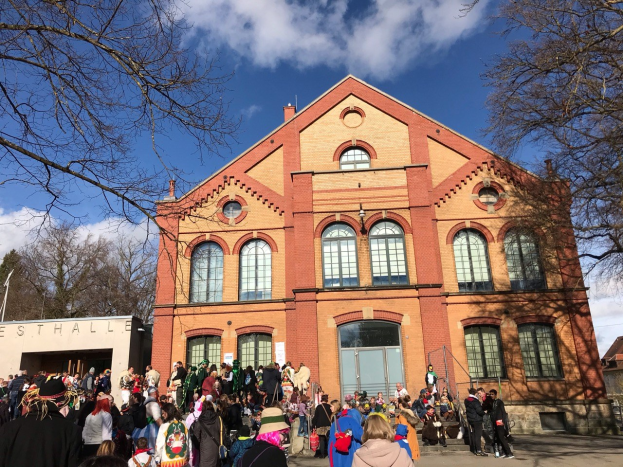 Eine große Gruppe von Menschen steht vor einem Gebäude mit Fenstern und Türen unter einem bewölkten Himmel, umgeben von Bäumen, einige tragen Mützen und Taschen, was auf ein Stadtfest hinweist.