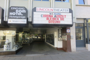 Das Lincoln Theater in Berlin, Deutschland, ein Gebäude mit Glasfenstern und -türen und einer Tafel mit Text darauf, das verschiedene Objekte im Inneren zeigt und so den Eindruck einer pulsierenden Stadtkulisse vermittelt.