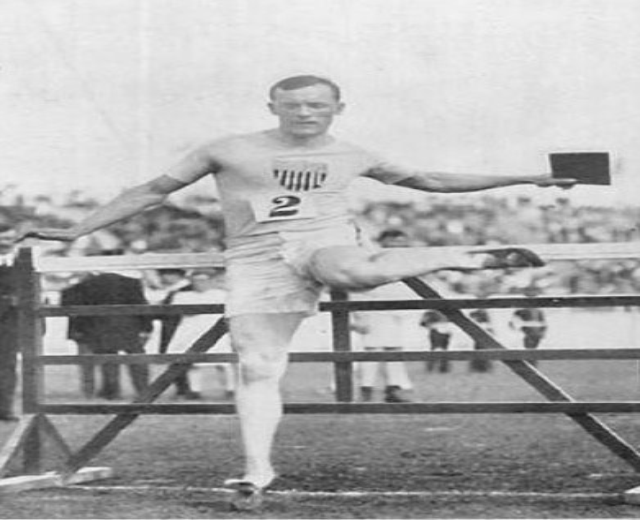 Schwarz-weiß-Foto eines Mannes in Sportkleidung, der über ein Hindernis springt, mit einer Menschenmenge und Himmel im Hintergrund.
