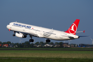 Ein Airbus A330-300 der Turkish Airlines startet vom Frankfurter Flughafen, mit Gras am Boden, Gebäuden und Bäumen im Hintergrund und Windmühlen in der Ferne unter einem klaren blauen Himmel.