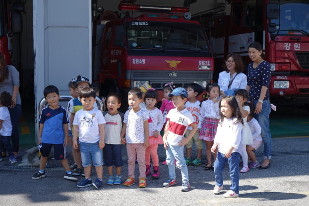 Eine Gruppe von Kindern vor einem Feuerwehrauto an einer Feuerwache, einige tragen Mützen, mit weiteren Feuerwehrautos im Hintergrund.