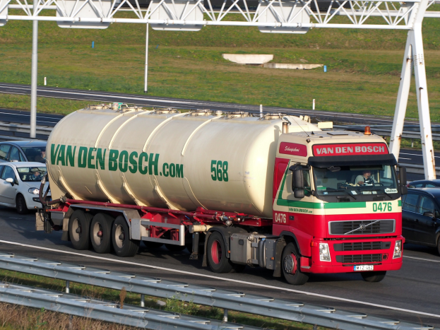 Ein großer weißer, roter und grüner Tanklastwagen fährt auf einer Autobahn neben anderen Fahrzeugen, mit einer Begrenzung am Straßenrand, Gras im Hintergrund und einer Eisenrahmenkonstruktion.