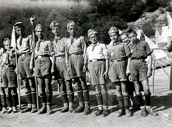 Schwarz-weiß-Foto einer Gruppe von Pfadfindern in Uniform, die Fahnen halten, mit Zelten und Bäumen im Hintergrund.
