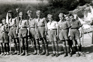 Schwarz-weiß-Foto einer Gruppe von Pfadfindern in Uniform, die Fahnen halten, mit Zelten und Bäumen im Hintergrund.