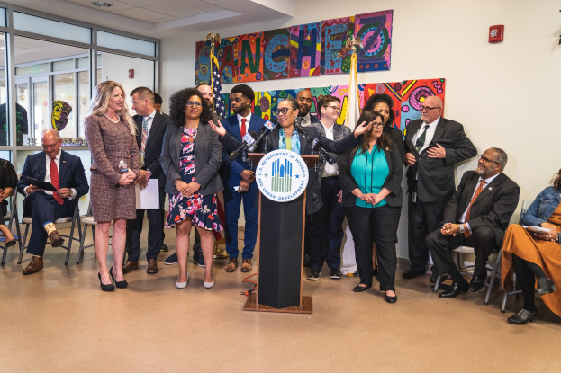 Gruppe von Menschen vor einem Rednerpult mit Mikrofon und einer Tafel mit der Aufschrift "Sancho-Stadtrat tritt dem Stadtrat bei", mit sitzenden und stehenden Teilnehmern, Fahnen, Feuerlöscher und Deckenleuchten.