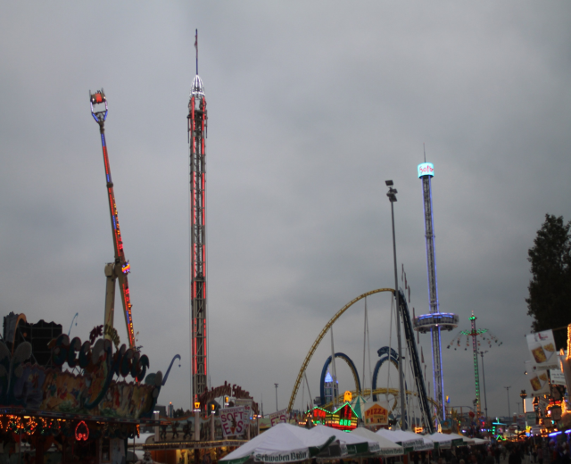 Menschenmenge bei nächtlichem Karneval mit beleuchteten Fahrgeschäften, Pfählen und Bäumen unter einem bewölkten Himmel.