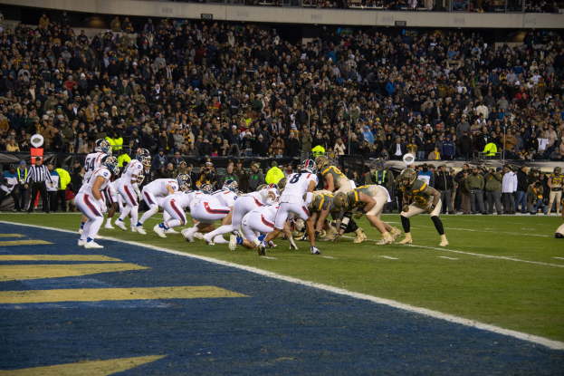 Ein Fußballspiel im Gange mit Spielern auf dem Boden in Sportkleidung und Helmen, von einem Stadion aus mit Zuschauern und Tafeln im Hintergrund betrachtet.