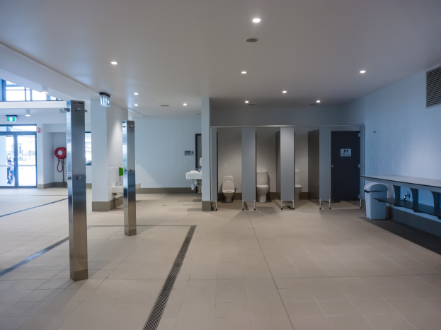 Große ßeffentliche Toilette mit mehreren Toiletten, tragenden Säulen, einem Mülleimer, einer Tür, einem Schild, einem Feuerlöscher und Deckenleuchten.