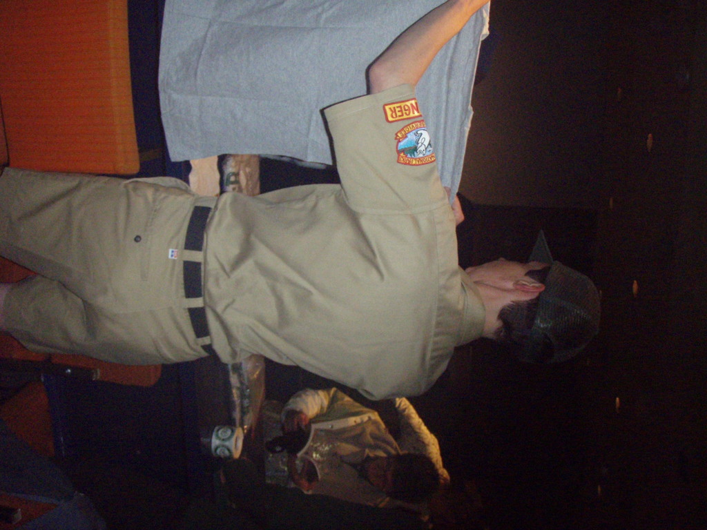 Ein Polizist hält ein T-Shirt in den Vordergrund, mit einem Jungen im Hintergrund, der ein Buch und einen Becher hält, der in der Nähe eines Schreibtischs und Lampen steht.