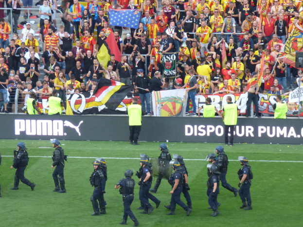 Eine Gruppe von Polizisten in Helmen und mit Waffen in der Hand läuft über ein Fußballfeld, im Hintergrund halten Menschen Fahnen, Schilder und Geländer.