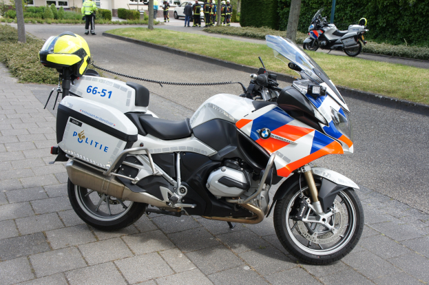 Polizeimotorrad am Straßenrand mit einer Gruppe von Menschen in bunten Kleidern und Helmen, Bäume, Pflanzen und Gebäude im Hintergrund.