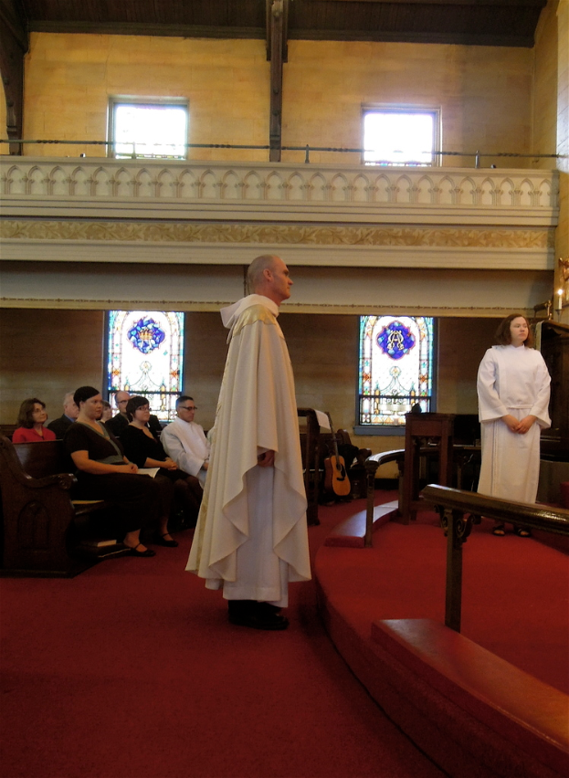 Eine Frau und ein Mann in weißen Kleidern stehen in einer Kirche, mit einem roten Teppich zu ihren Füßen, hölzernen Geländern, Kerzen, Bänken mit sitzenden Menschen und Musikinstrumenten im Hintergrund.