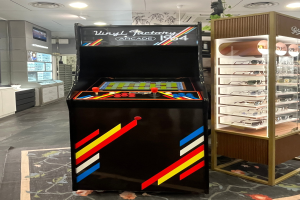Ein Geschäft mit einem zentralen Retro-Arcade-Automaten, einem rechten Gestell mit Gläsern und Hintergrund-Elementen wie Schränken, einem Fernseher und anderen Gegenständen, mit Deckenlampen, die für eine Retro-Atmosphäre sorgen.