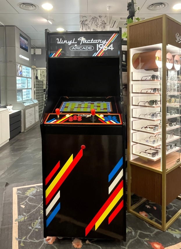 Ein Geschäft mit einem zentralen Retro-Arcade-Automaten, einem rechten Gestell mit Gläsern und Hintergrund-Elementen wie Schränken, einem Fernseher und anderen Gegenständen, mit Deckenlampen, die für eine Retro-Atmosphäre sorgen.