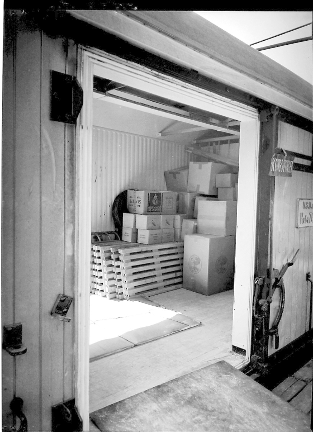 Schwarze und weiße Fotografie eines Metall-Shipping-Containers mit einer Tür auf der linken Seite, gefüllt mit ordentlich gestapelten Kartons.