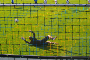 Ein Fussballer taucht ab, um einen Ball vor dem Tor zu halten, mit einem Netz im Vordergrund und einigen Menschen im Hintergrund, während mehrere Bälle auf dem Boden verstreut sind.