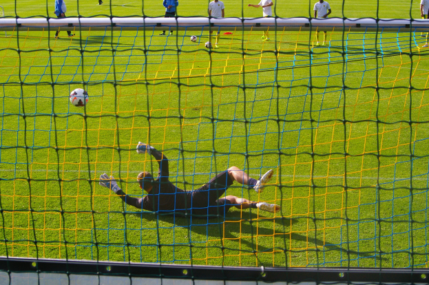Ein Fussballer taucht ab, um einen Ball vor dem Tor zu halten, mit einem Netz im Vordergrund und einigen Menschen im Hintergrund, während mehrere Bälle auf dem Boden verstreut sind.
