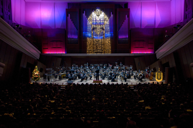 Großer Saal mit Publikum bei einem Konzert, mit Musikern auf der Bühne und sitzenden Personen, dekoriert mit einem Weihnachtsbaum, Lichtern und anderen Gegenständen.