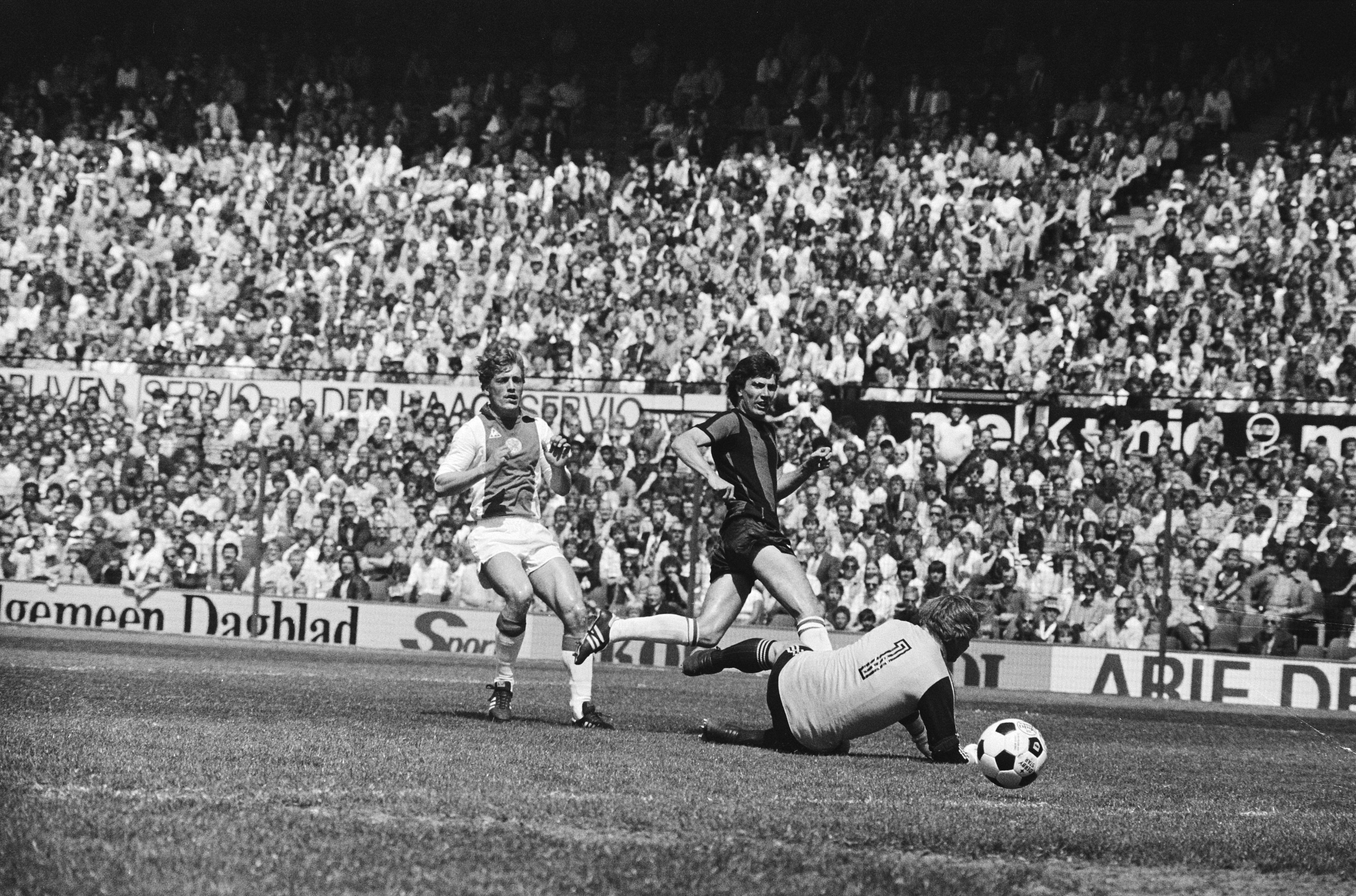 Schwarzes und weißes Foto von Fußballspielern im Einsatz auf einem Feld mit Zuschauern im Hintergrund und einer Bundesliga 1978-79 Ajax 1:1 Hannover 96-Banner.