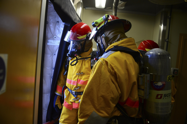 Feuerwehrleute in gelben Uniformen und roten Helmen stehen in einer Gruppe neben Zylindern und einer Tür, mit einer Wand und Licht im Hintergrund.