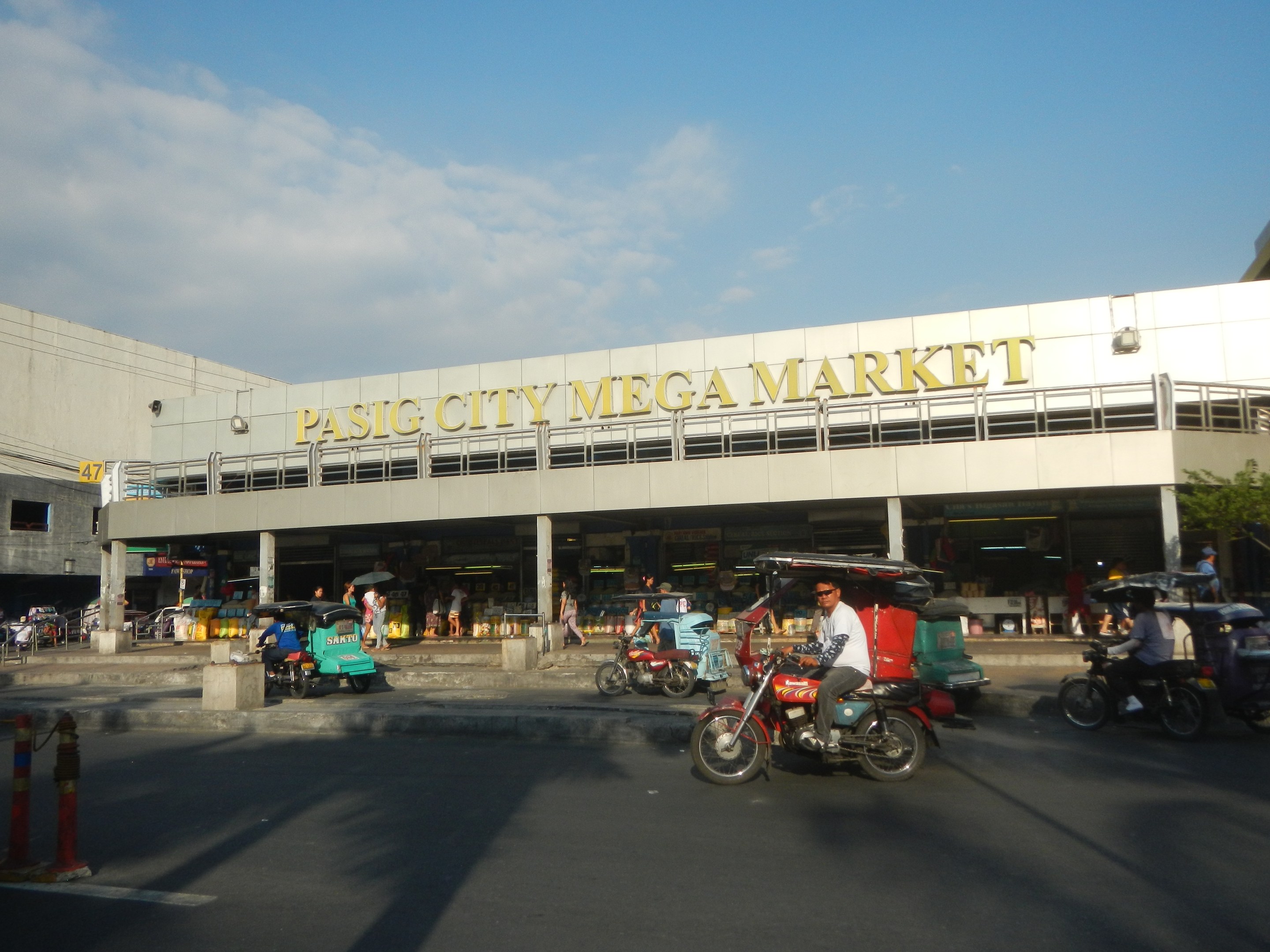 Eine belebte Straßenszene am Pasig City Mega Market mit Fahrzeugen und Fußgängern, mit einem Gebäude mit Text im Hintergrund unter einem bewölkten Himmel.