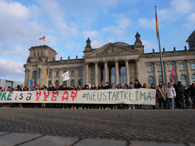 Gruppe von Menschen vor dem Reichstaggebäude in Berlin mit einer Fahne, auf der "Wir sind ein Menschenrecht" steht, zu sehen. Im Hintergrund sind die architektonischen Details des Gebäudes und Flaggen zu erkennen.