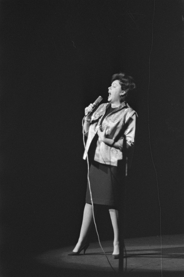 Eine Frau in einem Schwarz-Weiß-Outfit mit 1950er-Jahre-Frisur singt enthusiastisch in ein Mikrofon auf einer beleuchteten Bühne.