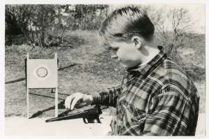 Ein junger Junge hält eine Waffe vor eine Zielscheibe mit Bäumen im Hintergrund, dargestellt in Schwarz-Weiß.