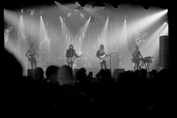 Ein Schwarz-Weiß-Bild eines Musikkonzerts mit vier Künstlern auf der Bühne: zwei Gitarristen und eine Sängerin, mit Zuschauern unten und Lichtern oben.