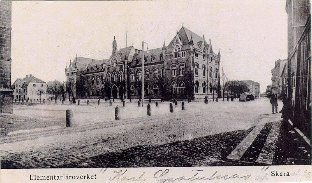 Ein Schwarz-Weiß-Foto einer Stadtstraße aus den frühen 1900er-Jahren, das Gebäude, Bäume, Pfähle, Menschen und Fahrzeuge zeigt, mit dem Text "Skara-Schule in den frühen 1900er-Jahren" unten.