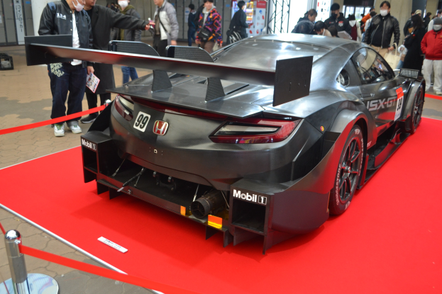 Ein roter und weißer Honda NSX GT3 Rennauto auf einer Indoor-Veranstaltung ausgestellt, umgeben von Menschen in bunten Kleidern und einigen mit Masken, mit einem roten Teppich auf der Seite und Anzeigetafeln im Hintergrund.