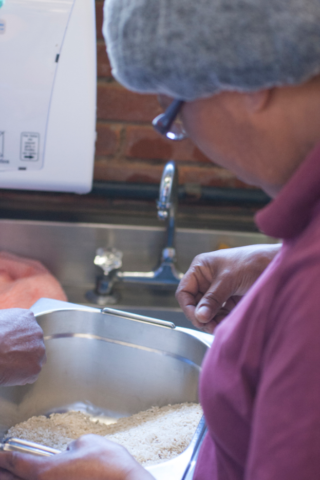 Eine Frau mit Brille und T-Shirt hält einen Stahlschüssel mit Reis neben einem Waschbecken mit Wasserhähnen, steht neben einer Ziegelwand, mit einer Maschine in der oberen linken Ecke.