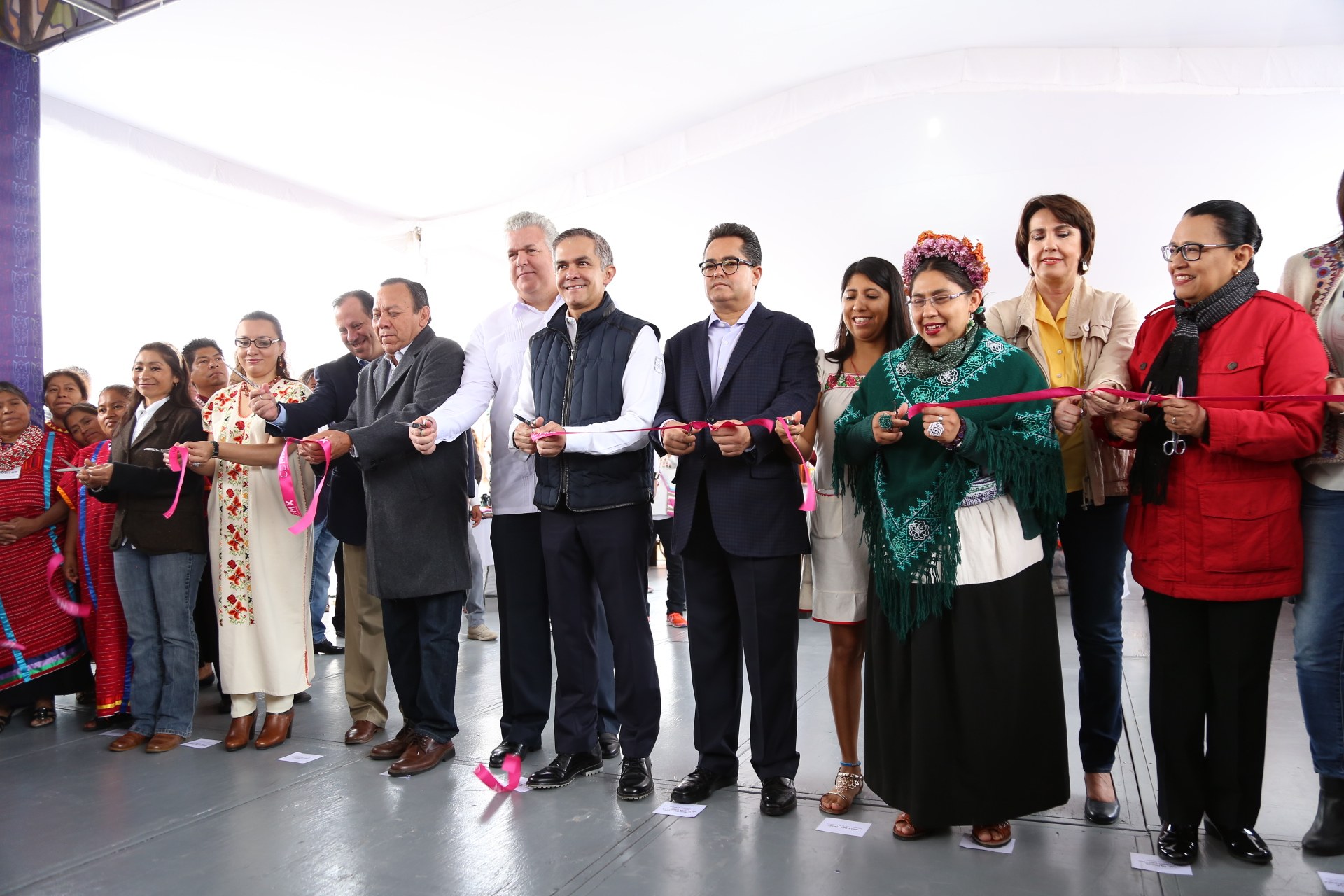 Eine Gruppe von Menschen bei einer Scherenschnitte-Zeremonie, mit einer Säule links, verstreuten Papieren auf dem Boden und einem weißen Tuch im Hintergrund.