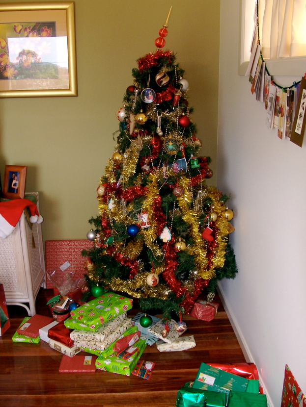 Ein geschmückter Weihnachtsbaum mit Geschenken darunter, ein Foto Rahmen an der linken Wand und ein Vorhang am rechten Fenster.