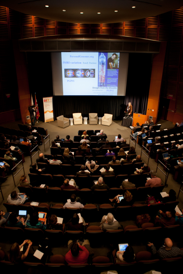 Ein vollbesetztes Auditorium mit sitzenden Menschen, die Gegenstände halten, beleuchtet von vielen Lichtern, mit Fahnen links, einem Podium rechts und einem Werbeplakat links.