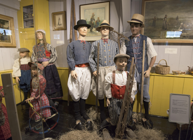 Eine Gruppe von Schaufensterpuppen in historischer Kleidung steht zusammen in einem Raum mit trockenem Gras auf dem Boden, einem Textbrett, an der Wand montierten Fotorahmen, einem Schrank mit Gegenständen und einem Korb und anderen Gegenständen auf einer Plattform, wahrscheinlich im Nationalmuseum für die Geschichte und Kultur der Vereinigten Staaten.