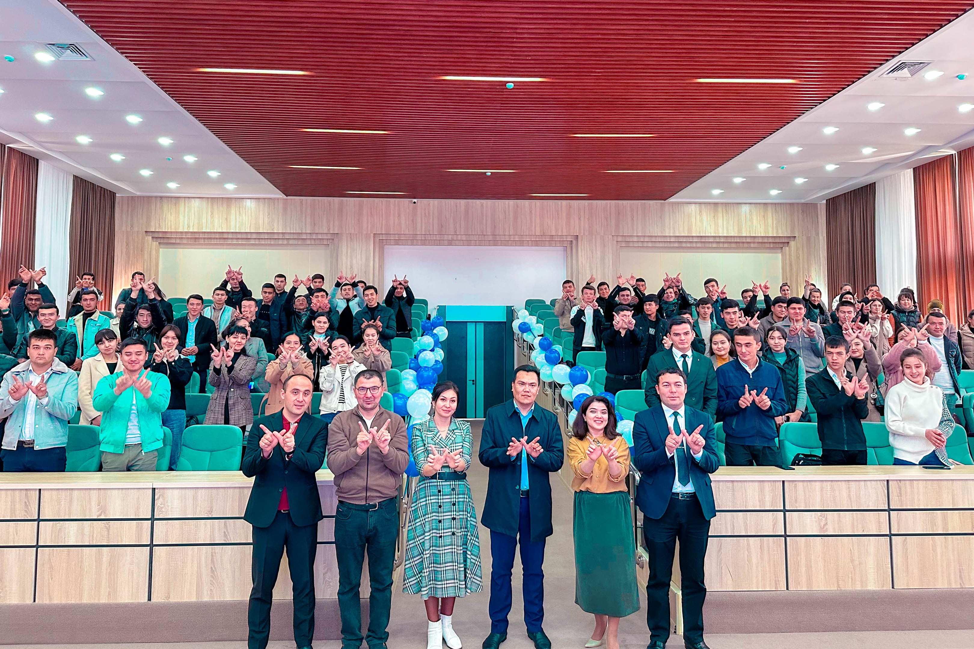 Eine Gruppe von Menschen steht vor einer Menge, einige klatschen, mit Stühlen, Tischen, Ballons, Vorhängen und einer Wand im Hintergrund und Lampen an der Decke, bei der Eröffnungsfeier einer neuen Schule.