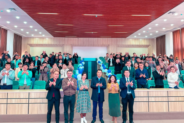 Eine Gruppe von Menschen steht vor einer Menge, einige klatschen, mit Stühlen, Tischen, Ballons, Vorhängen und einer Wand im Hintergrund und Lampen an der Decke, bei der Eröffnungsfeier einer neuen Schule.