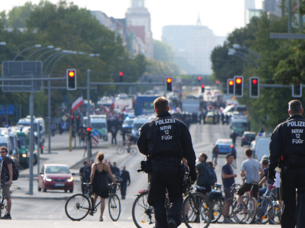 Zwei Polizisten neben einer Gruppe von Radfahrern auf einer Straße mit Fahrzeugen, Verkehrszeichen, Bäumen, Gebäuden und einem klaren blauen Himmel im Hintergrund.