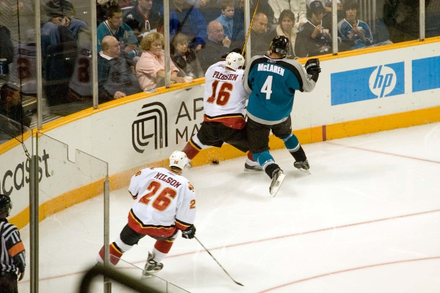 Ein Mann in einem sechzehn Nummern-Jersey, Helm, Shorts und Schuhen steht neben einem anderen Mann in einer blauen Jacke, Brille, Helm und Hose in der Nähe eines Fensters, mit einem Publikum, das oben sitzt und ein Spiel schaut, einem Mann in einem sechsundzwanzig Nummern-Jersey, der unten einen Stock hält, einem Schiedsrichter hinter Glas in der linken unteren Ecke und einer Werbetafel auf der rechten Seite.