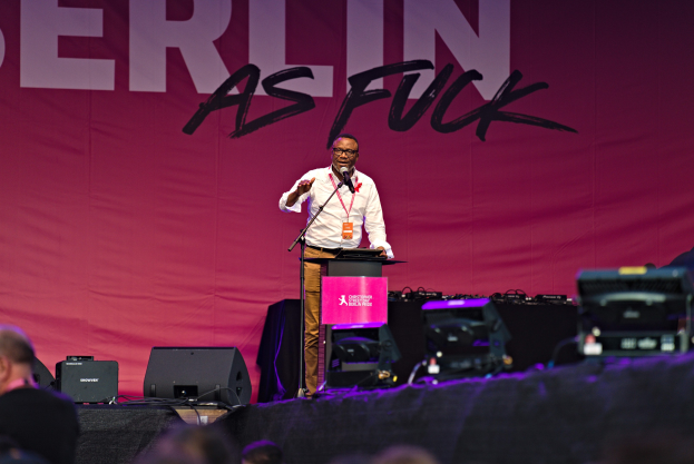 Ein Mann in einem weißen Hemd und braunen Hosen steht auf einer Bühne und hält ein Mikrofon in der Hand, während er zu einer Menge spricht. Im Hintergrund ist ein pinkfarbenes Banner mit der Aufschrift 'Berlin as Fuck' zu sehen.