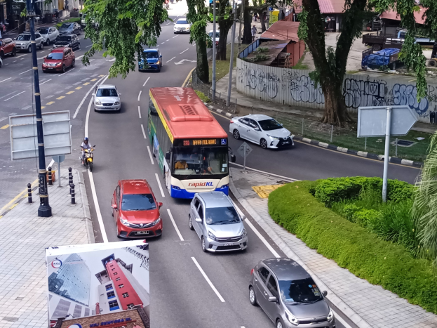 Eine belebte Stadtstraße mit Autos, Bussen, Motorrädern, Straßenlaternen, Bäumen und Gebäuden, einschließlich einer Wand mit sichtbarem Text.
