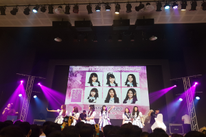 Eine Gruppe von Menschen auf einer hell erleuchteten Bühne, die vor einem Publikum performs, mit einem großen Bildschirm im Hintergrund, der Bilder der K-Pop-Gruppe BK48 anzeigt.