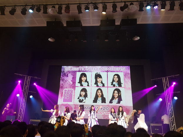 Eine Gruppe von Menschen auf einer hell erleuchteten Bühne, die vor einem Publikum performs, mit einem großen Bildschirm im Hintergrund, der Bilder der K-Pop-Gruppe BK48 anzeigt.