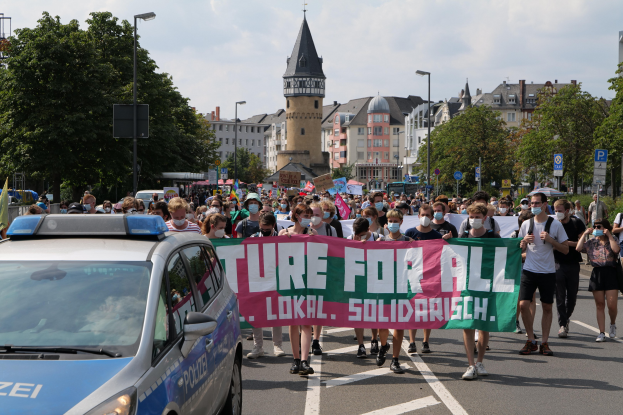 Eine große Gruppe von Menschen marschiert mit einem "Lokal Solidarität"-Schild die Straße entlang, mit einem Polizeiwagen neben ihnen geparkt, Bäumen, Laternen, Schildern, Gebäuden und einem bewölkten Himmel im Hintergrund.