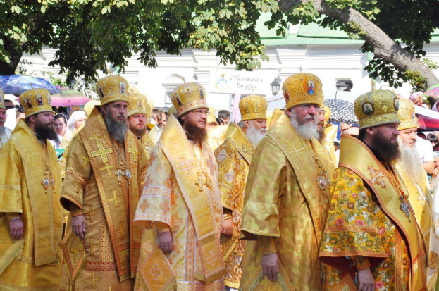 Eine Gruppe von Priestern in gelben Roben und Hüten, die eine Straße entlanggehen und von Menschen mit Schirmen umgeben sind, mit einem Gebäude und einem Baum im Hintergrund.