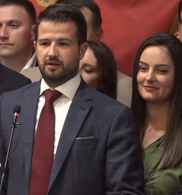 Ein Mann im Anzug spricht an einem Rednerpult mit einem Mikrofon zu einer Menge hinter einem Banner, wahrscheinlich über ein Regierungssozialmedienverbot.