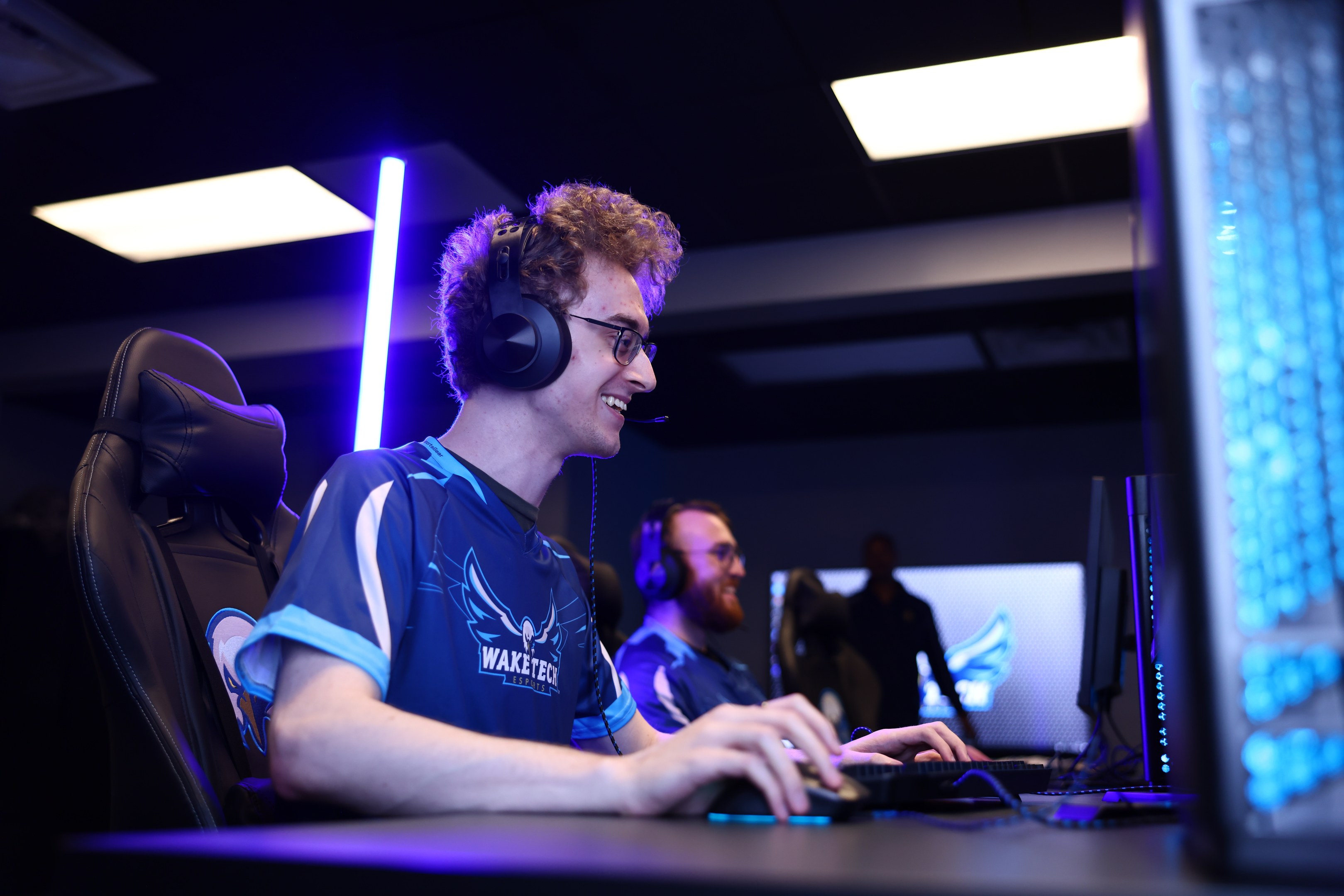 Ein Mann trägt einen Headset und Brille und sitzt an einem Schreibtisch mit einem Computerbildschirm, einer Tastatur und einer Maus, während er in einem Raum mit einer anderen Person und Deckenlampen spielt.