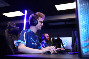 Ein Mann trägt einen Headset und Brille und sitzt an einem Schreibtisch mit einem Computerbildschirm, einer Tastatur und einer Maus, während er in einem Raum mit einer anderen Person und Deckenlampen spielt.